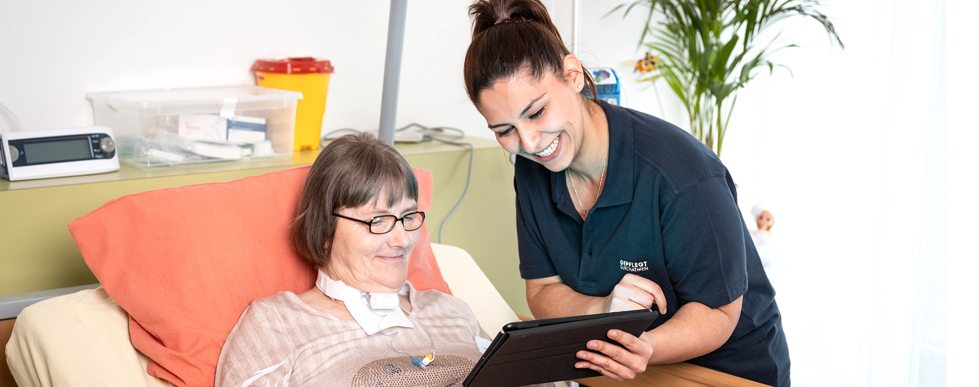 Patientin sitzt im Krankenbett. Eine weibliche Pflegekraft zeigt ihr etwas vornübergebeugt auf einem Laptop.
