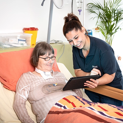 Patientin sitzt im Krankenbett. Eine weibliche Pflegekraft zeigt ihr etwas vornübergebeugt auf einem Laptop.